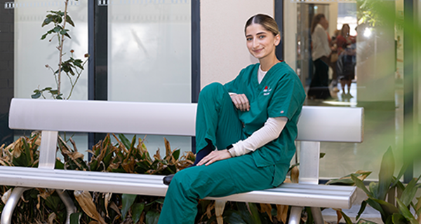 Student in scrubs sitting on a silver bench outside of a hospital