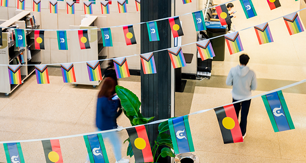 Atrium adorned with colorful Aboriginal, Torres Strait Islander, and rainbow flags, as two people walk beneath festive bunting.