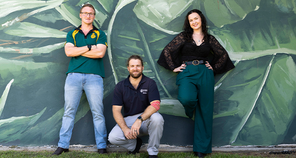 Three students posing in front of a wall that features a green leaf mural