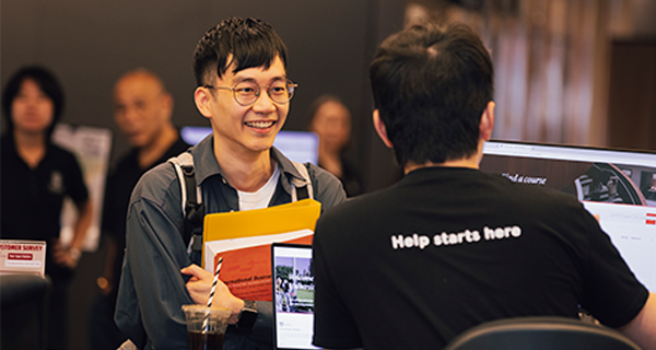 A student holding a book and folder speaking with someone that has 'Help starts here' on the back of their shirt.