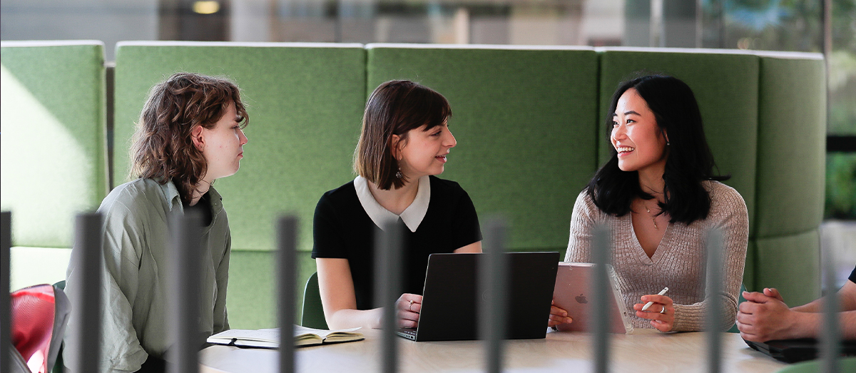 Three students talking to each other while sitting at a desk with an open book, laptop and iPad in front of them