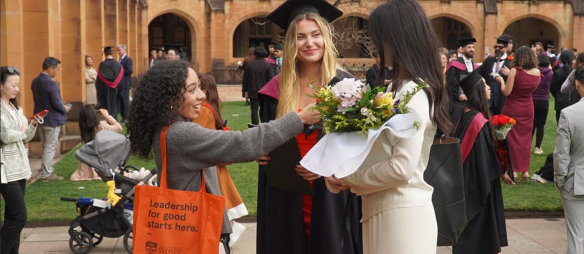 A student holding a microphone with a tote bag that says 'Leadership for good starts here.' The student is holding a microphone towards a graduate holding flowers, with another graduate looking over