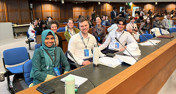 Students sitting at a long desk in a lecture hall, smiling at the camera