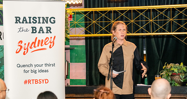 A person on stage speaking with a microphone in front of them with a 'Raising the Bar' banner beside them