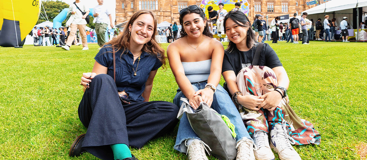 Three students sitting on a lawn with stalls in the background, smiling.