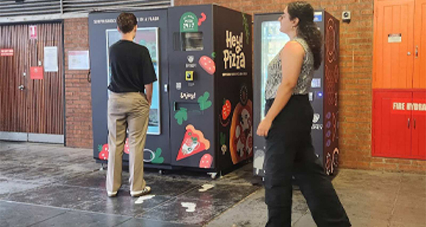 A student standing in front of a pizza vending machine with another student in frame walking past